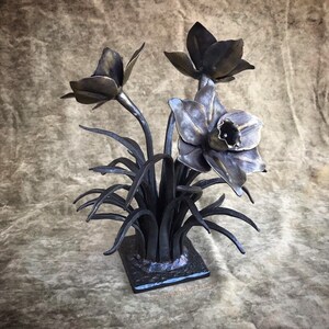 Könnte beinhalten: Eine schwarze Metallsculptur eines Straußes Narzissen. Die Blumen sind aus Metall und haben ein realistisches Aussehen. Die Skulptur steht auf einem kleinen quadratischen Sockel.