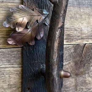 May include: A rustic bronze door handle with a branch-like grip and a decorative plate featuring oak leaves. The handle is mounted on a wooden door, showcasing a natural, handcrafted design.