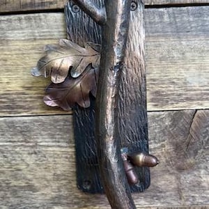 May include: Bronze-toned metal door pull with a rustic design. The pull features a textured, bark-like background, a central branch, oak leaves, and an acorn. The hardware is mounted on a wooden surface.