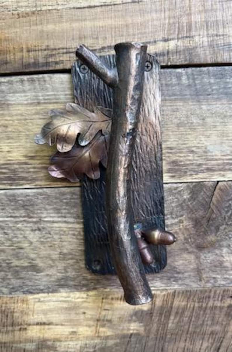 May include: Bronze-toned metal door pull with a rustic design. The pull features a textured, bark-like background, a central branch, oak leaves, and an acorn. The hardware is mounted on a wooden surface.
