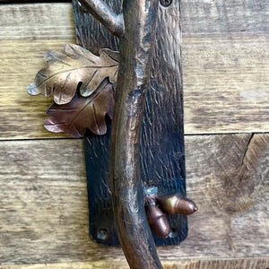 May include: A bronze-toned metal door handle with a tree branch design. The handle features a textured tree trunk, oak leaves, and acorns. The handle is mounted on a rectangular plate with a screw.