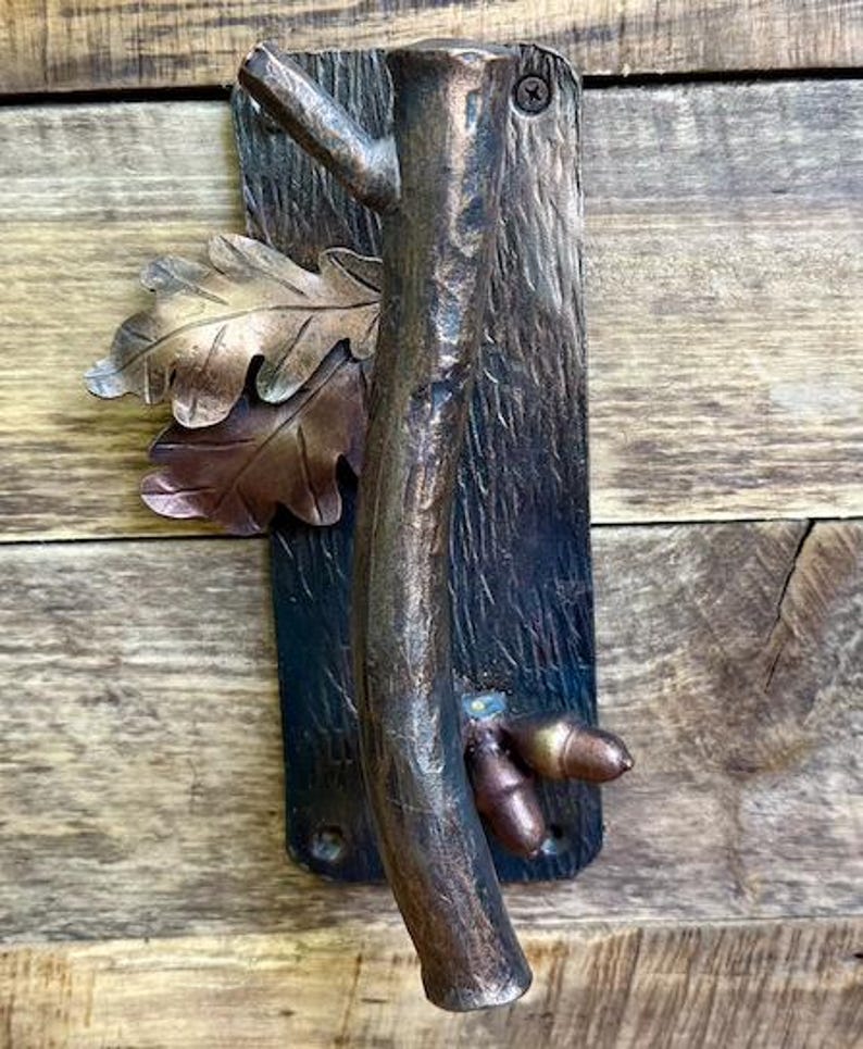 May include: A bronze-toned metal door handle with a tree branch design. The handle features a textured tree trunk, oak leaves, and acorns. The handle is mounted on a rectangular plate with a screw.