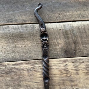 May include: A dark brown, hand-carved wooden wand with a curved top and a detailed face carving. The wand features a spiral design on the lower portion and a crescent moon detail near the top.