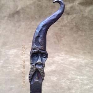 May include: Hand-forged metal wizard hook with a crescent moon and face design. The hook is dark with a curved top and the text "Burnt Whisker Forge".