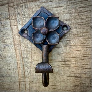 Puede incluir: Un gancho de pared decorativo de metal bronce oscuro. El gancho presenta una placa trasera en forma de diamante con un diseño floral y un gancho curvo. El gancho está montado sobre una superficie de madera.