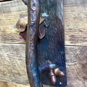 May include: A rustic bronze door handle with a tree branch design. The handle features a textured branch, leaves, and acorns. The handle is mounted on a dark wooden plate with a single screw.