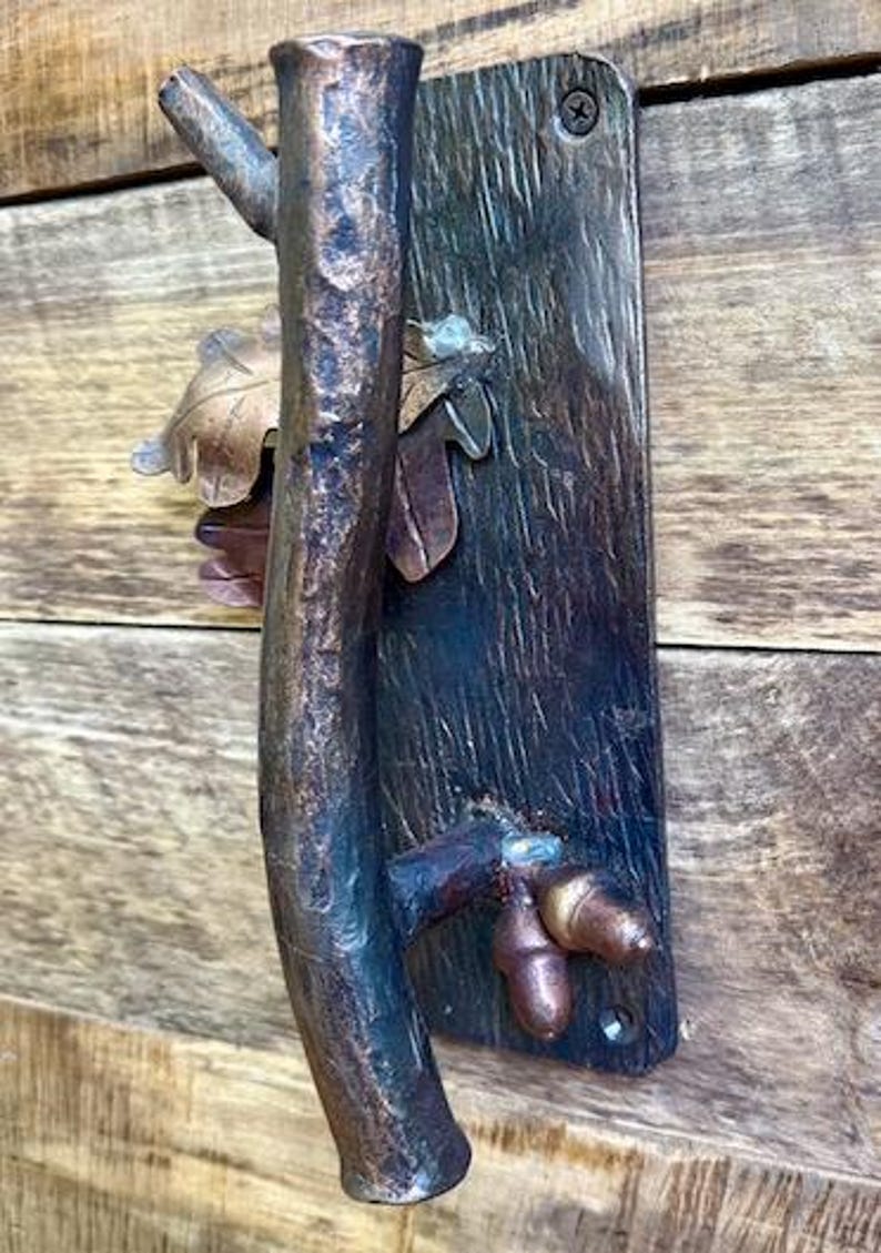 May include: A rustic bronze door handle with a tree branch design. The handle features a textured branch, leaves, and acorns. The handle is mounted on a dark wooden plate with a single screw.