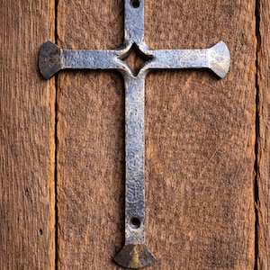 May include: A handcrafted metal cross with a rustic, aged appearance. The cross is mounted on a weathered wooden surface, showcasing its detailed design and texture. The cross has a diamond-shaped cutout at the intersection.