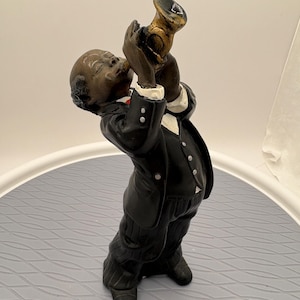 May include: A figurine of a musician playing a gold-colored trumpet. The figure wears a black suit jacket with white buttons, a white shirt, and black pinstriped trousers. The musician has dark skin and is captured in a standing pose with eyes closed.