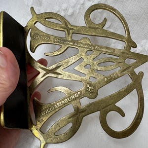 May include: A vintage brass bookend with an ornate, openwork design. The bookend features the text "Williamsburg" and "The Colonial Williamsburg Foundation" along with the year "1977". The bookend has a black backing.