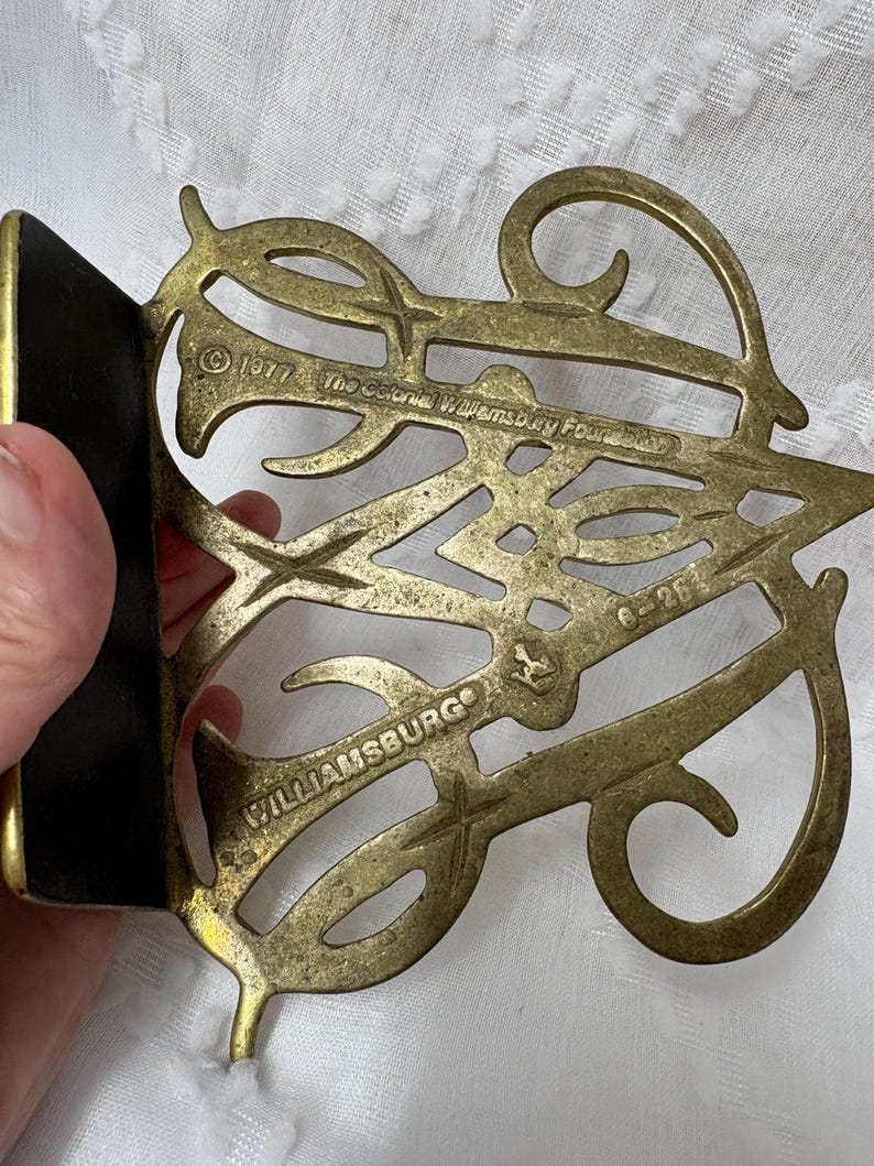 May include: A vintage brass bookend with an ornate, openwork design. The bookend features the text "Williamsburg" and "The Colonial Williamsburg Foundation" along with the year "1977". The bookend has a black backing.