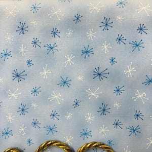 May include: Light blue fabric with a repeating pattern of white and blue starburst designs. The fabric is displayed against a gold-colored, ornate metal frame, creating a decorative presentation.