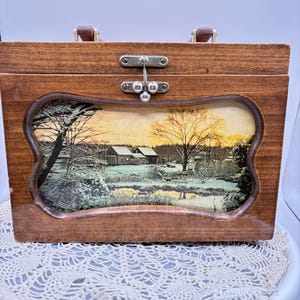 May include: A rectangular wooden box with a hinged lid and a metal latch closure. The front panel features a recessed, wavy-edged window displaying a printed winter landscape scene with snow-covered houses, bare trees, and a golden sunset sky.
