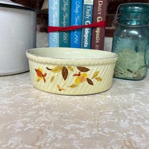 May include: A round, pale yellow ceramic bowl featuring a diagonal ribbed texture and a floral pattern with brown, orange, and yellow leaves and blossoms. The rim has a subtle metallic silver edge, and the bowl sits in front of several books.