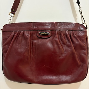 May include: A burgundy leather shoulder bag with a gathered design. The bag has a long, adjustable strap and a small, rectangular emblem on the front. The bag's color is a deep, rich shade of red-brown, and it appears to be vintage.