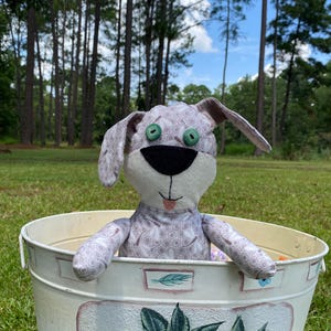 May include: A plush gray and white dog toy with green button eyes sits in a white metal bucket with a floral design. The bucket is sitting on a grassy surface.