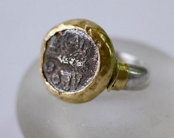 May include: A silver ring with a gold band. The ring features a silver coin with a design of a bird and inscription.