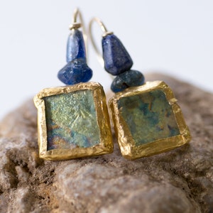 Könnte beinhalten: Ein Paar goldene Ohrringe mit quadratischen blauen und grünen Glasstücken. Die Ohrringe haben eine goldene Drahtschlaufe und eine blaue Glasperle.