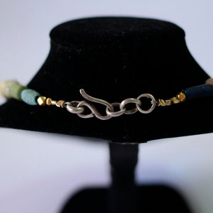 May include: A close-up of a silver clasp on a multi-colored beaded necklace. The necklace features green, blue, and gold beads.