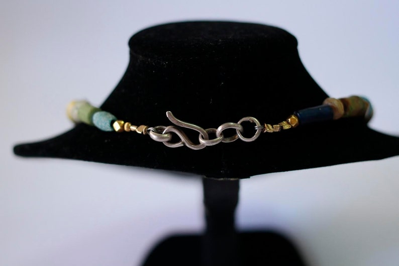 May include: A close-up of a silver clasp on a multi-colored beaded necklace. The necklace features green, blue, and gold beads.