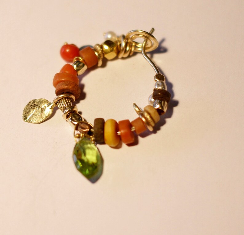 K&ouml;nnte beinhalten: Goldene Creolen mit einer Auswahl an bunten Perlen, darunter orange, gelb, braun und gr&uuml;n. Die Ohrringe haben einen Blattanh&auml;nger und einen kleinen gr&uuml;nen Edelstein.