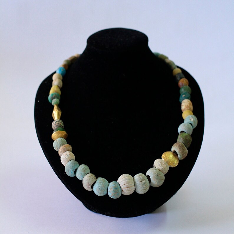 May include: A necklace made of blue, green, and gold beads. The beads are strung on a black cord and are displayed on a black velvet stand.