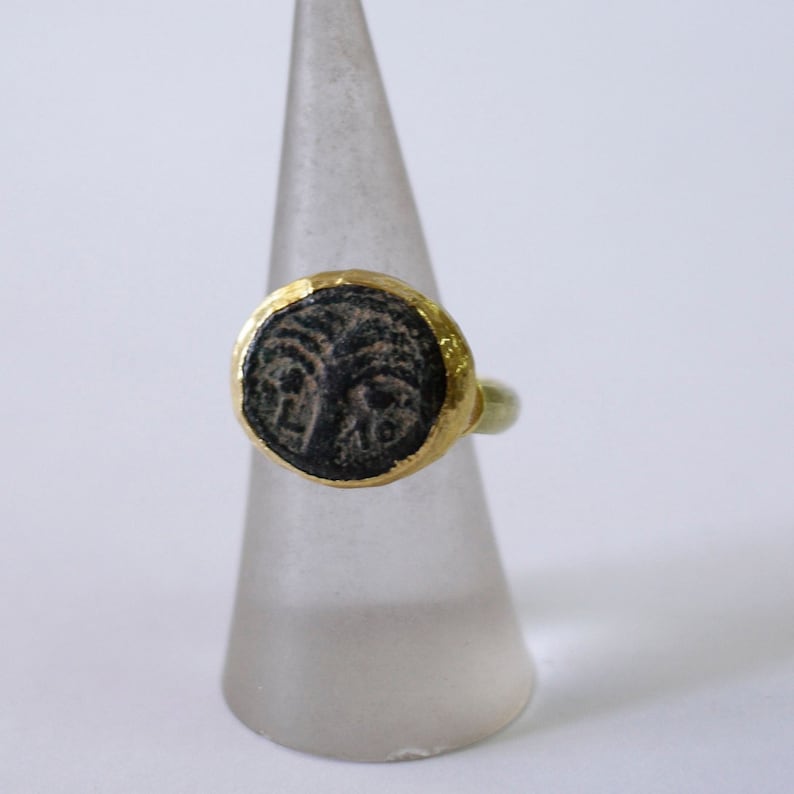May include: A gold ring with a black oval stone featuring an intricate design. The ring is displayed on a white cone-shaped stand.