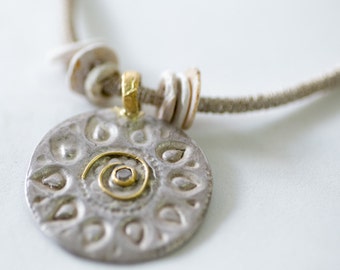 May include: A silver pendant necklace with a circular design featuring a small diamond in the center. The pendant is surrounded by a gold swirl and a series of teardrop shapes. The necklace is made of a brown cord with white beads.