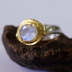 Könnte beinhalten: Ein Silber- und Goldring mit einem runden Mondstein, der in einer goldenen Fassung sitzt. Der Ring liegt auf einem braunen Stein.