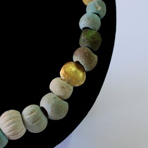 May include: A close-up of a necklace made of various colored beads, including blue, green, yellow, and gold. The beads are strung on a black cord and are arranged in a gradient pattern.