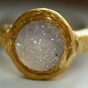 May include: A gold ring with a white iridescent stone set in the center. The stone is made of many small pieces of white material that shimmer in the light.