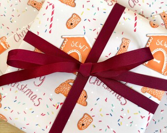 Puede incluir: Un papel de regalo blanco con un patrón repetido de casas de jengibre, bastones de caramelo y perros. Las casas están decoradas con el texto "SCWT" y las palabras "Feliz Navidad" están impresas en el papel de regalo. Una cinta roja está atada alrededor del papel de regalo.