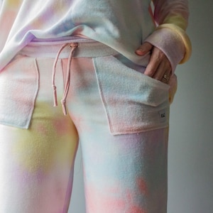 May include: Tie-dye sweatpants with a pink drawstring and a small patch on the right pocket. The sweatpants are a light pink, blue, and yellow color.