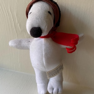 May include: A white plush Snoopy toy wearing a brown aviator cap and a red scarf. The toy has black eyes and a black nose. A tag on the toy reads "FOR CHILDREN AGES 3 AND UP SURFACE WASH ONLY ALL NEW CONTENTS: POLYESTER FIBERS REG. NO. PA-7059 (HK)"