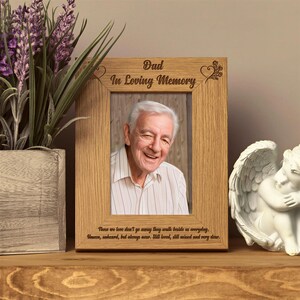 a wooden picture frame with a photo of an elderly man, placed on a wooden shelf next to a white ceramic angel figurine and a potted plant.