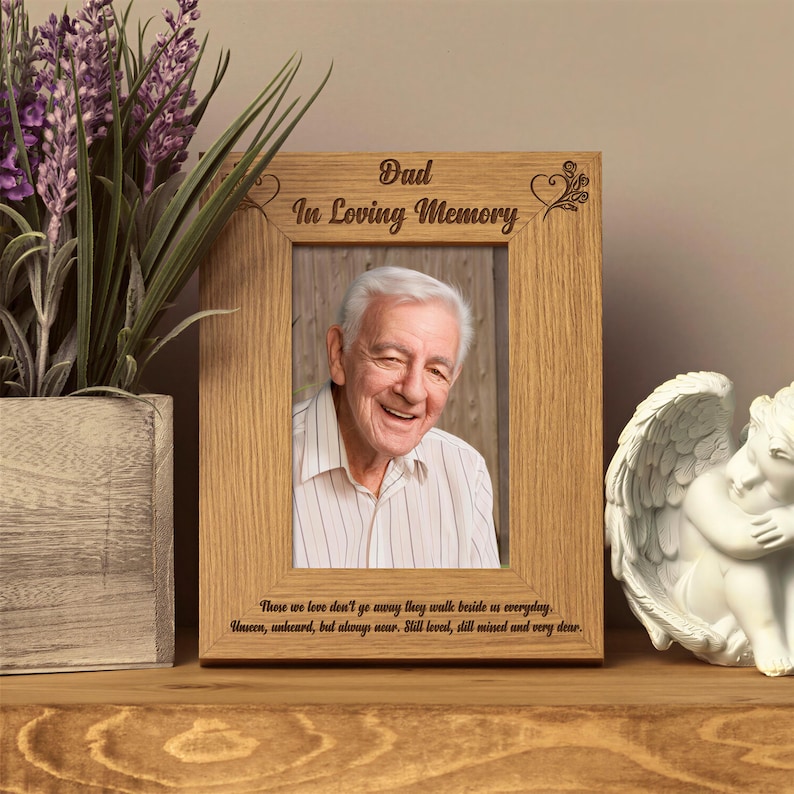 a wooden picture frame with a photo of an elderly man, placed on a wooden shelf next to a white ceramic angel figurine and a potted plant.