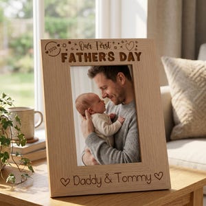 A wooden picture frame with a photo of a man holding a baby, and the text "Our First Father's Day" engraved on it.