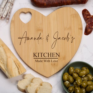 May include: A heart-shaped bamboo cutting board with the text "Amanda & Jacob's Kitchen Made With Love" engraved on it. The board has a small heart-shaped cutout in the top.