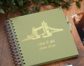 May include: A green photo album with a gold foil design of the Tower Bridge in London. The album has a spiral binding and a ribbon closure. The text "Natalie & Jack London 2024" is printed in gold foil on the front cover.
