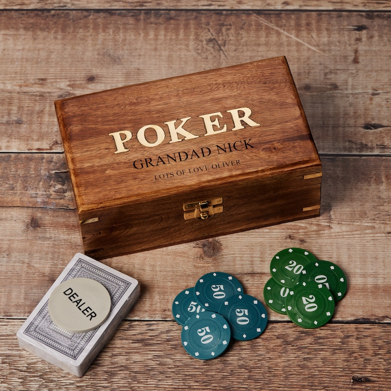 May include: A wooden box with a hinged lid and a brass latch. The box is engraved with the words "POKER GRANDAD NICK LOTS OF LOVE OLIVER". The box is sitting on a wooden surface with green poker chips and a deck of cards.