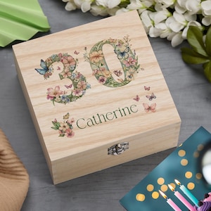 Puede incluir: Una caja de madera de color claro con el número "30" y el nombre "Catherine" impresos en la tapa. Los números y el nombre están decorados con diseños florales y de mariposas. La caja tiene un cierre metálico.