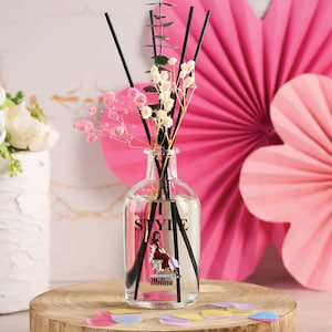 May include: A clear glass bottle diffuser with black reeds and a label that says "I Style" with a woman in a red dress sitting on a chair. The label also says "Melissa". The bottle is surrounded by pink and white flowers.