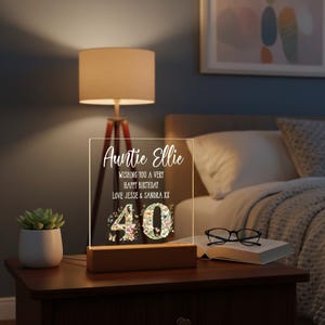 May include: A clear acrylic LED night light with a wooden base. The light is personalized with the text "Auntie Ellie" and a birthday message. The number "40" is decorated with floral designs. The lamp is on a dark wooden nightstand.