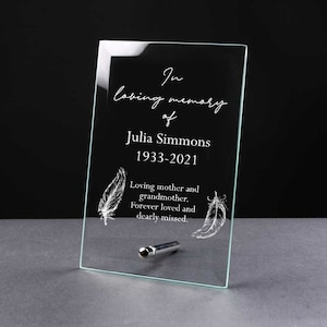 May include: A glass memorial plaque with a white inscription that reads "In loving memory of Julia Simmons 1933-2021 Loving mother and grandmother. Forever loved and dearly missed." The plaque has two etched feather designs.