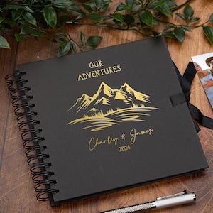 May include: Black photo album with a gold mountain range design and the text "Our Adventures" and "Charley & James 2024".
