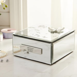 May include: A mirrored jewellery box with a butterfly accent on the lid. The box has a rectangular shape and a small plaque with text. A pearl bracelet and earrings are next to the box. The box is on a white surface.