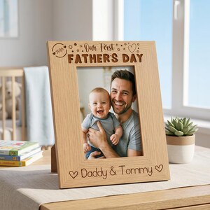 A wooden picture frame with a photo of a smiling man holding a baby, placed on a table with a potted plant and books in the background.