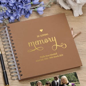 May include: A brown leather-bound memory book with gold lettering. The cover reads "In Loving Memory" with a heart symbol and the name "Phyllis Rose Marshall" with dates "22nd July 1936 - 09th March 2024".