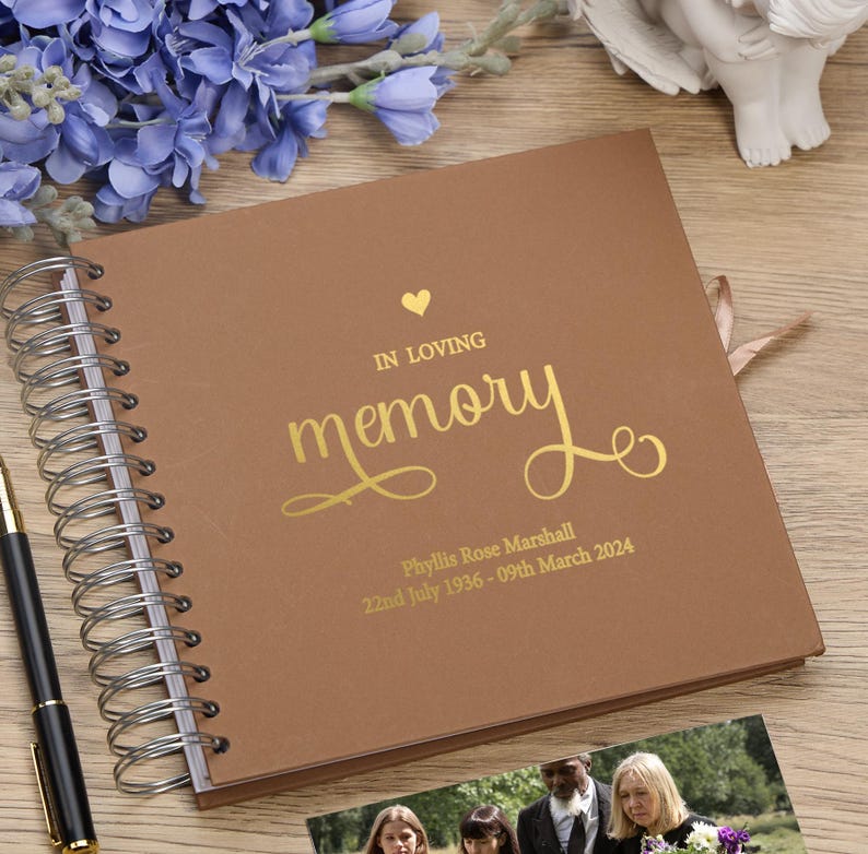 May include: A brown leather-bound memory book with gold lettering. The cover reads "In Loving Memory" with a heart symbol and the name "Phyllis Rose Marshall" with dates "22nd July 1936 - 09th March 2024".