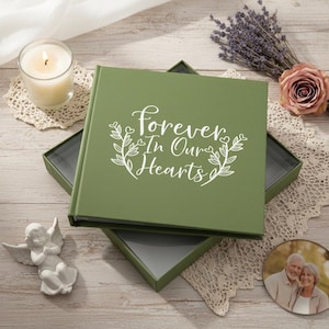 Puede incluir: Un álbum de fotos verde oliva con las palabras "Forever In Our Hearts" en escritura blanca. El álbum está abierto en una caja a juego. Una vela encendida, una figura de ángel y una foto también están en la imagen.
